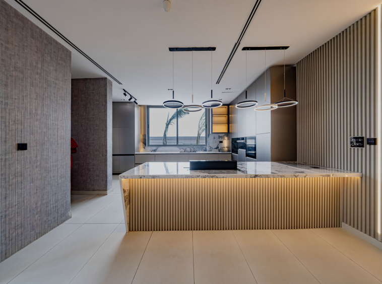 Modern kitchen interior with marble island, circular pendant lights, and a wooden slat wall along the right; large window shows palm trees outside.