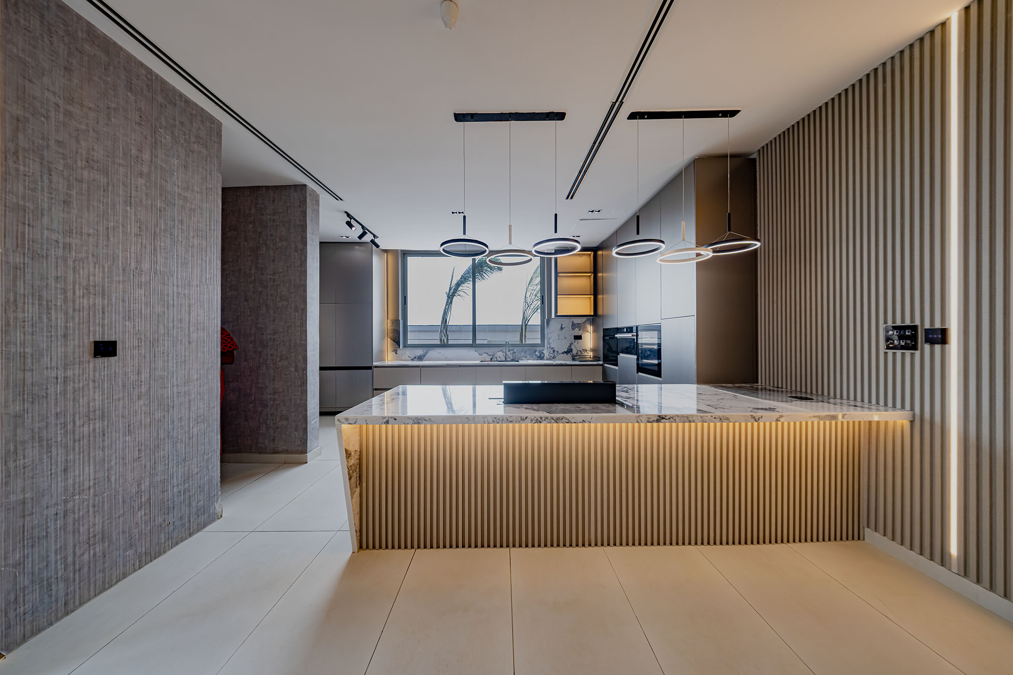 Modern kitchen interior with marble island, circular pendant lights, and a wooden slat wall along the right; large window shows palm trees outside.