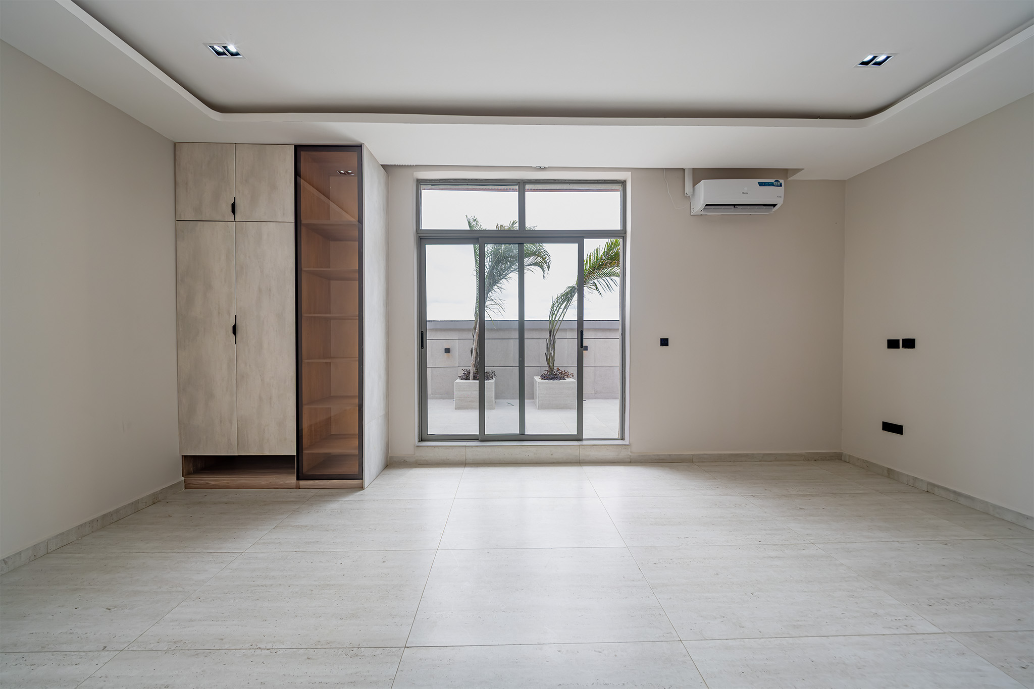 Bright empty room with a large glass sliding door to a patio and two potted palms outside; built-in cabinet on the left.