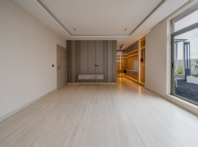 Bright open-plan room with beige walls, a wood-paneled feature wall, and built-in shelving illuminated by warm LEDs; large sliding glass door to an outdoor patio on the right.