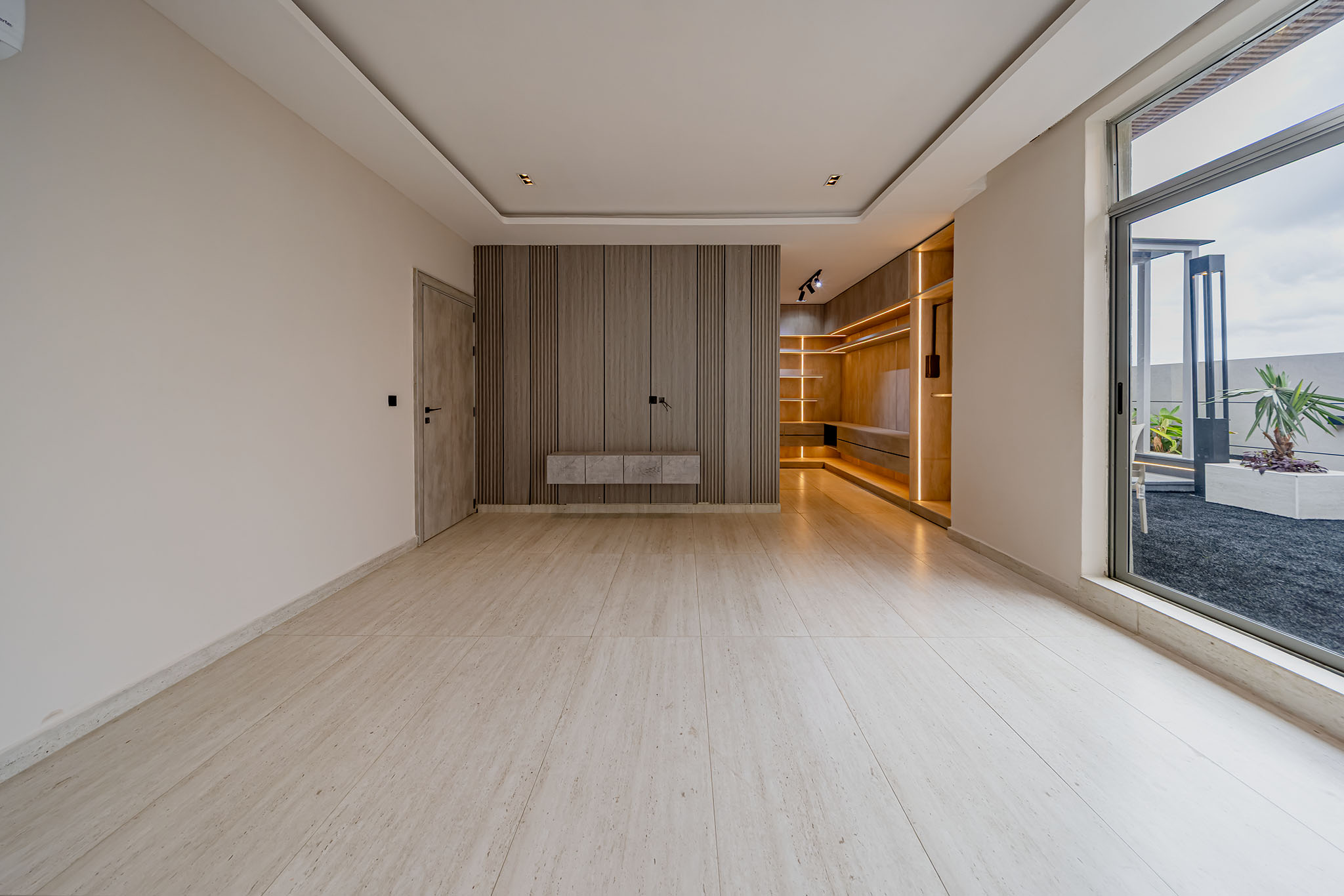 Bright open-plan room with beige walls, a wood-paneled feature wall, and built-in shelving illuminated by warm LEDs; large sliding glass door to an outdoor patio on the right.