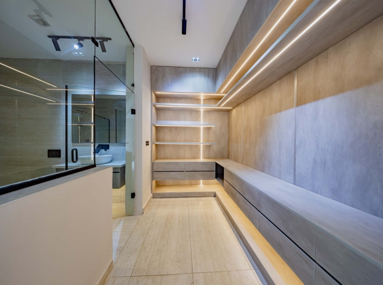 Modern walk-in closet with built-in shelves and drawers and warm wall lighting; glass bathroom wall on the left.
