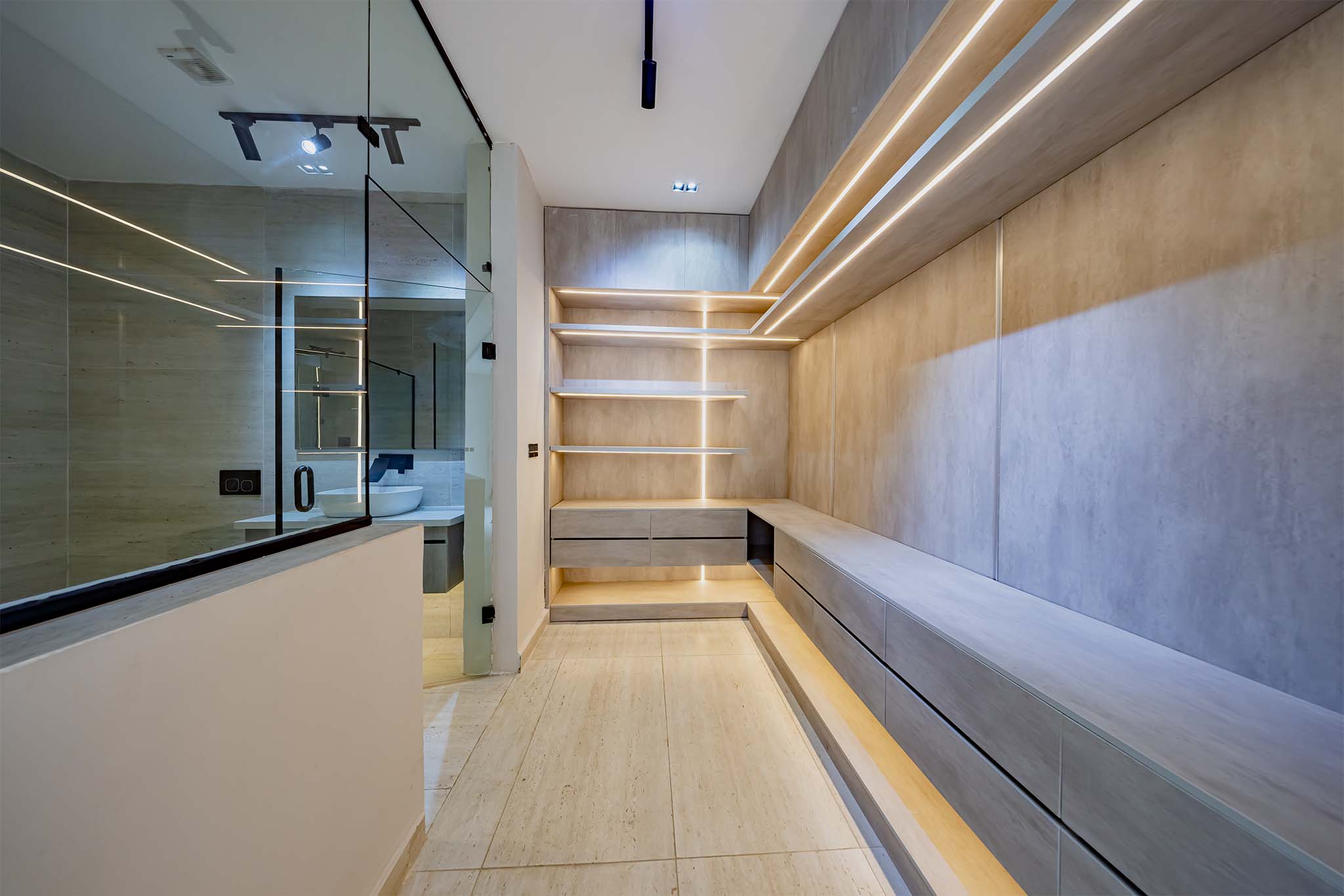 Modern walk-in closet with built-in shelves and drawers and warm wall lighting; glass bathroom wall on the left.