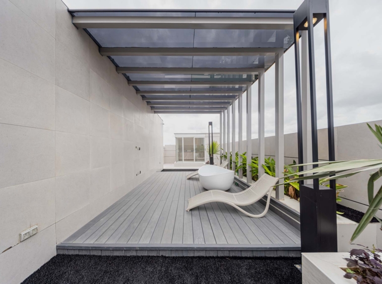 Modern outdoor patio with gray wooden decking, white curved lounge chairs, and a glass-roofed pergola supported by black metal beams along a beige tiled wall. Lush green plants line the edge.