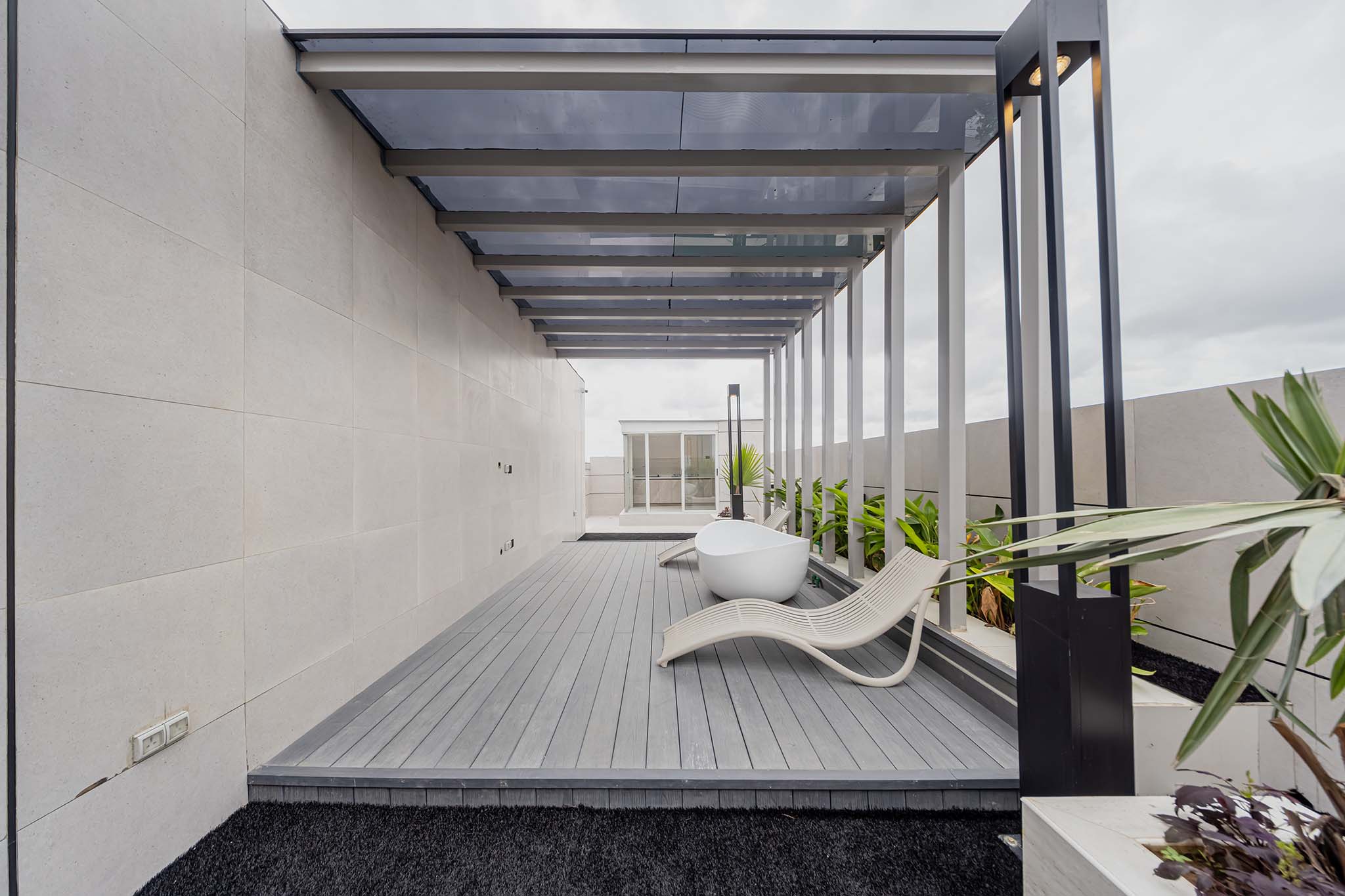 Modern outdoor patio with gray wooden decking, white curved lounge chairs, and a glass-roofed pergola supported by black metal beams along a beige tiled wall. Lush green plants line the edge.