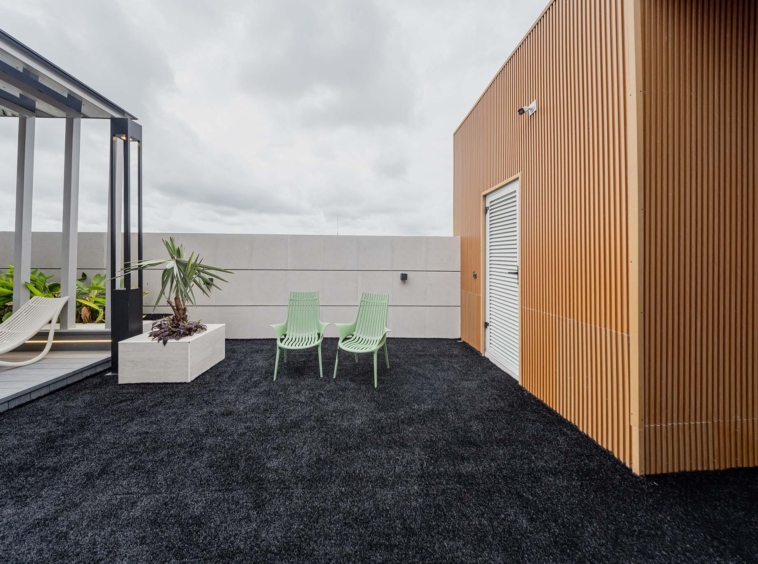 Rooftop terrace with black gravel, mint-green chairs, white planters, and a tall orange wood-panel wall on the right side.