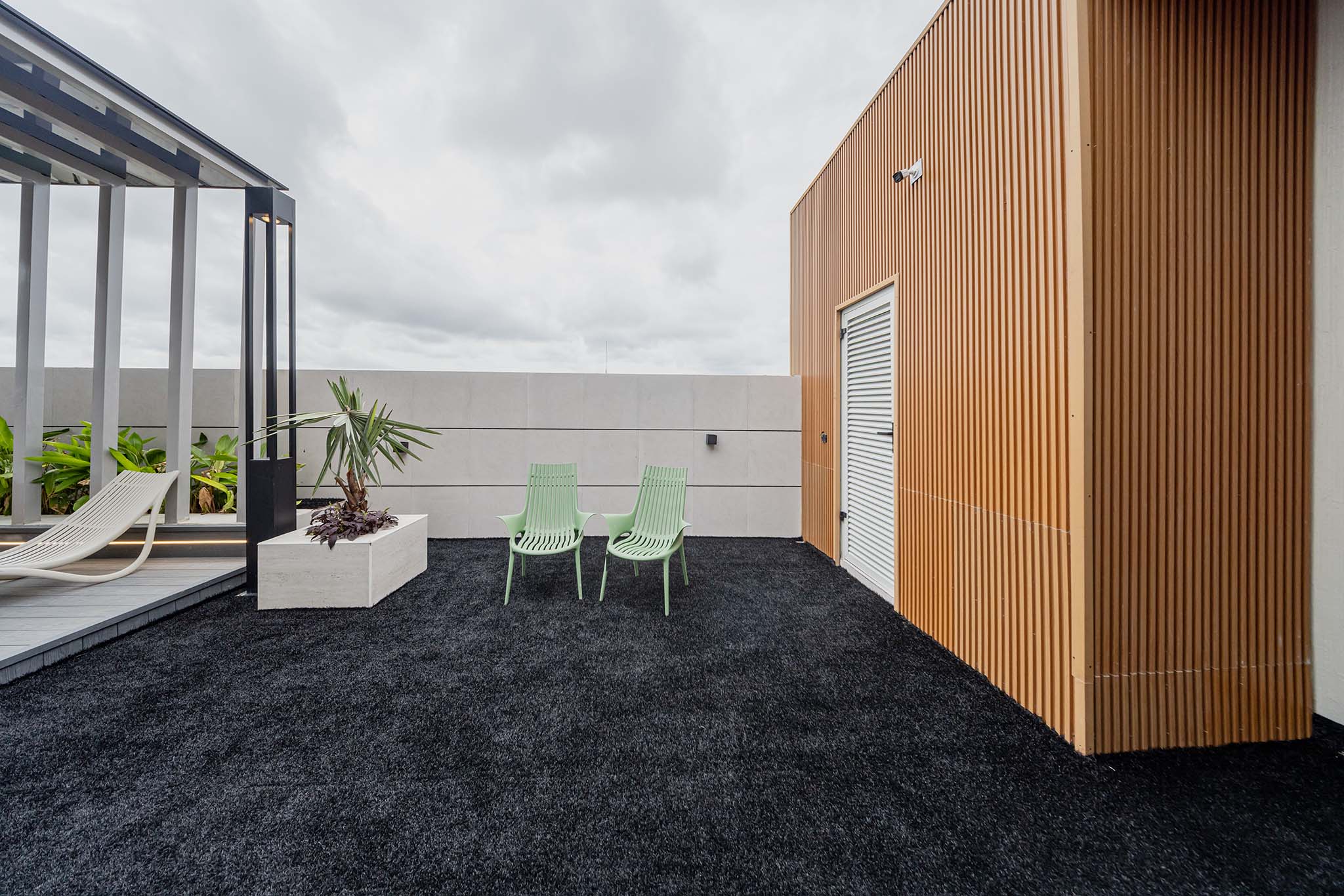 Rooftop terrace with black gravel, mint-green chairs, white planters, and a tall orange wood-panel wall on the right side.