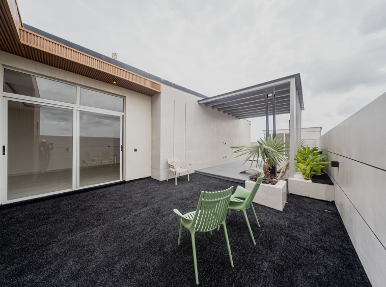 Modern rooftop patio with white walls, sliding glass doors, and a wooden slatted overhang; green plastic chairs and planters outline the space.