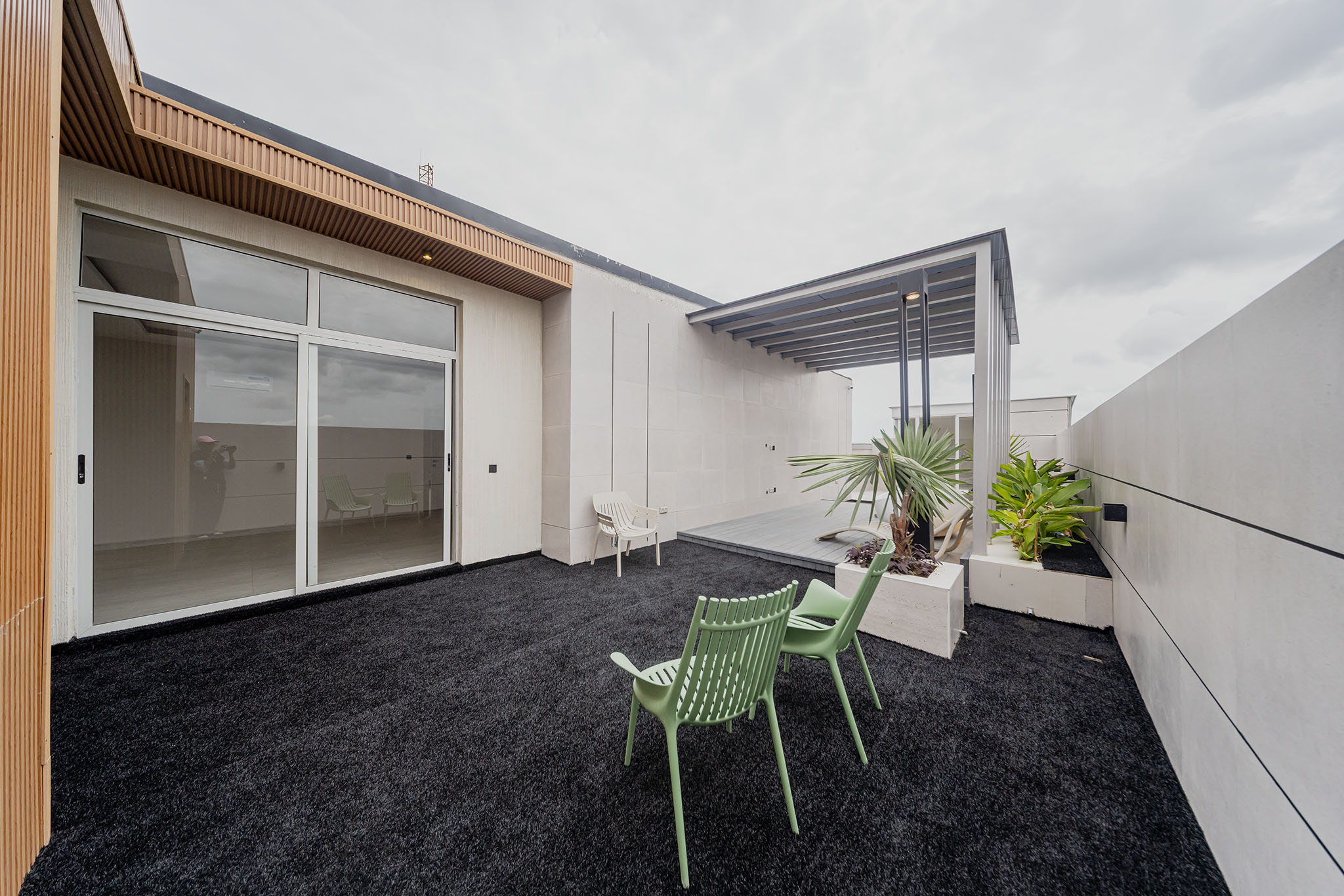 Modern rooftop patio with white walls, sliding glass doors, and a wooden slatted overhang; green plastic chairs and planters outline the space.