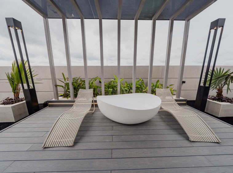 Modern rooftop lounge with curved cream loungers around a white oval tub beneath a metal pergola, surrounded by plants and a cloudy sky.