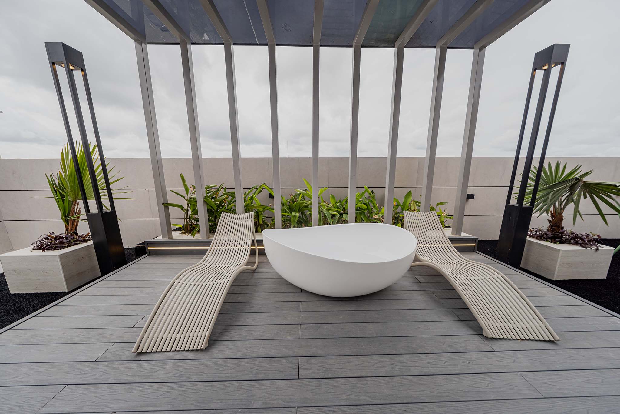 Modern rooftop lounge with curved cream loungers around a white oval tub beneath a metal pergola, surrounded by plants and a cloudy sky.