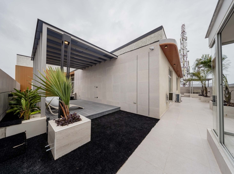 Modern rooftop courtyard with white tile flooring, black gravel, and large planters under a metal canopy attached to a beige building.