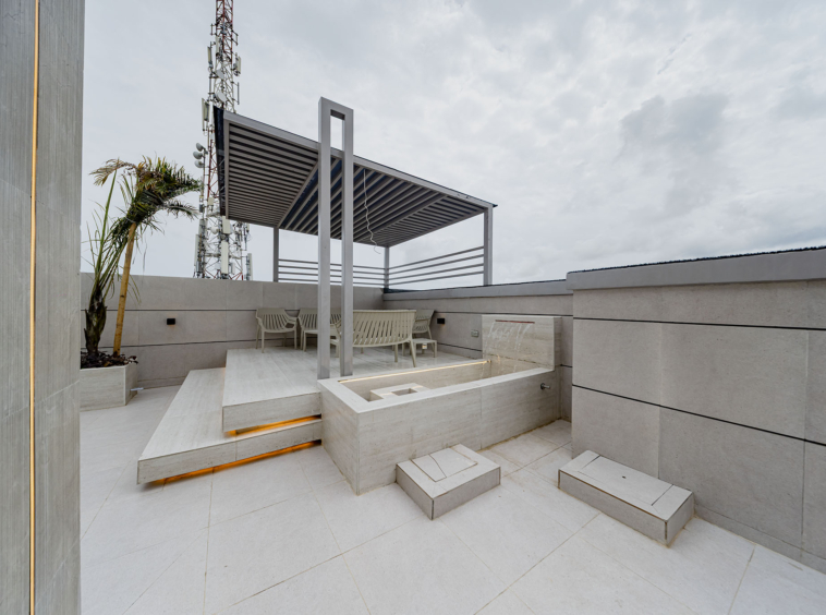 Modern rooftop patio with a metal slatted pergola, beige seating, and a palm plant against light gray walls, cloudy sky above.