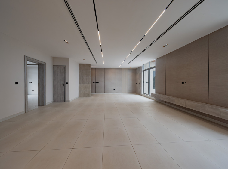 Bright, empty modern interior hallway with beige tiled floor, wood panel walls, and long skylight-like ceiling lighting on the right side of the room.