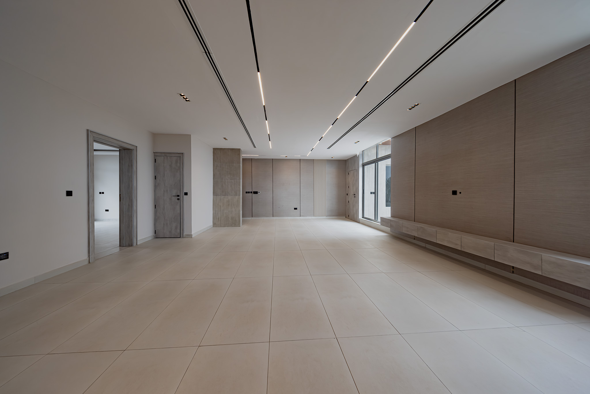 Bright, empty modern interior hallway with beige tiled floor, wood panel walls, and long skylight-like ceiling lighting on the right side of the room.