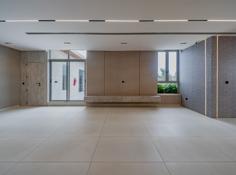 Modern minimalist lobby with light tiled floor, wood-paneled wall, and large windows showing greenery outside under a white ceiling.