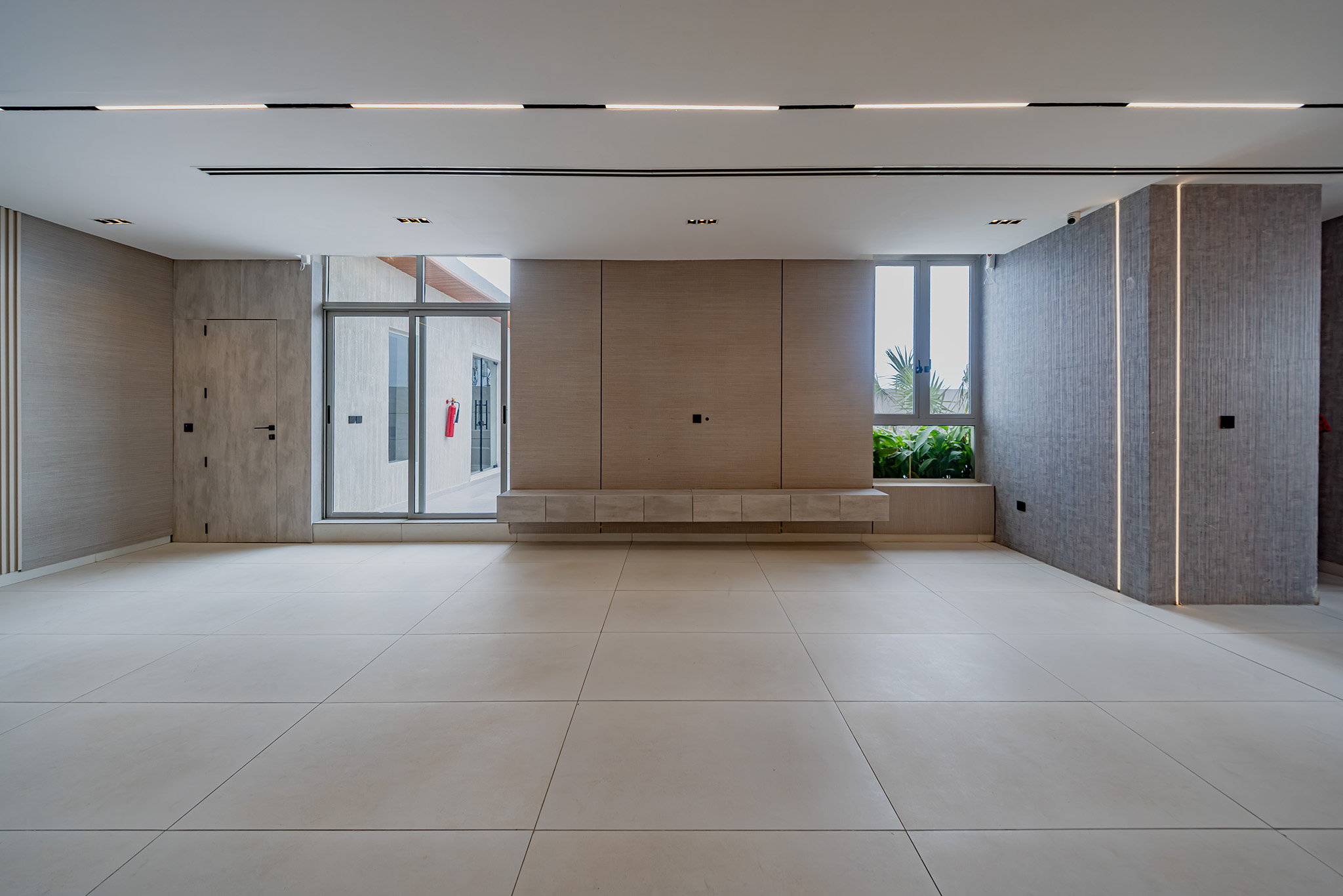 Modern minimalist lobby with light tiled floor, wood-paneled wall, and large windows showing greenery outside under a white ceiling.
