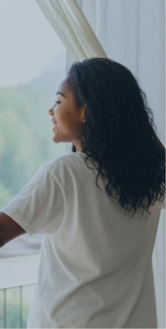 Woman with dark curly hair in a white shirt standing at a window, looking outside at a green landscape.